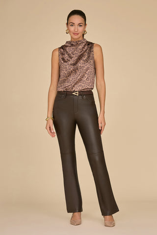 A person stands against a beige background wearing the Mara Silk Top in a leopard print and black leather pants. They have hoop earrings, a bracelet, and pointed nude shoes. Posing confidently with hands relaxed by their sides, they exude effortless style.