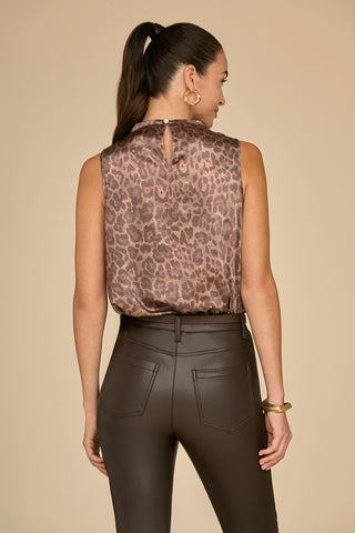 A woman with long hair in a ponytail is seen from behind, wearing the Mara Silk Top in leopard print and high-waisted black leather pants. Her gold hoop earrings and bracelets add a touch of elegance against the beige background.