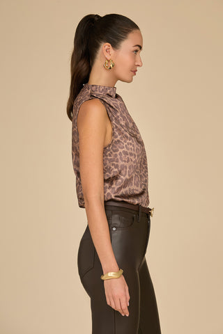 A woman with long dark hair in a ponytail is wearing the Mara Silk Top in a leopard print along with dark pants. She accessorizes with gold hoop earrings and a gold bracelet, standing in profile against a plain beige background and exuding effortless elegance.