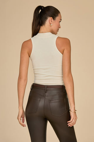 A woman with long dark hair in a ponytail faces away, highlighting her slim silhouette in the ultra-soft Talia Ribbed Tank - Pearl and dark high-waisted pants as she stands against a beige background.