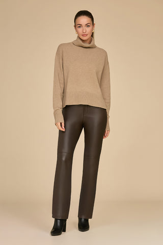 A person stands against a beige background, wearing a stylish Gia Turtleneck Sweater in cafe color, paired with dark brown leather pants and black ankle boots. Their hair is tied back, and they maintain a neutral expression.