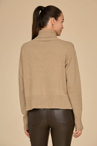 A person with a high ponytail is facing away, wearing a Cafe Gia Turtleneck Sweater and dark pants. The neutral background complements the outfit's earthy tones.