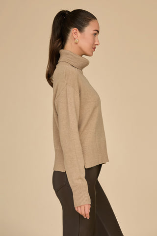 A person with long dark hair in a ponytail is wearing a Cafe-colored Gia Turtleneck Sweater and dark pants. They are facing to the right, standing against a plain beige background.