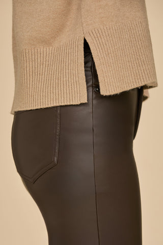 A close-up of a person wearing a Cafe Gia Turtleneck Sweater with ribbed details paired with dark brown leather pants. The focus is on the side view of the fabric textures and clothing fit against a neutral background.