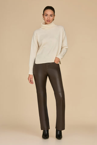 A woman stands against a neutral backdrop, wearing the Gia Turtleneck Sweater in Pearl paired with brown leather pants. Her hair is tied back, one hand resting in her pocket. Black ankle boots finish off her chic look.