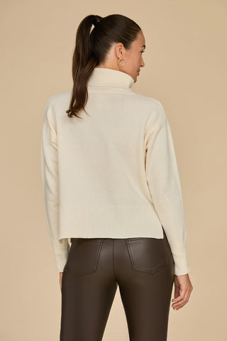 A woman with a high ponytail faces away from the camera, draped in an oversized Gia Turtleneck Sweater in Pearl and dark brown leather pants. The ensemble is set against a soft beige backdrop.