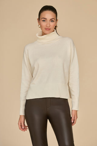 A person wearing an oversized Gia Turtleneck Sweater in Pearl and dark pants poses against a beige background. They have their hair pulled back and are looking slightly off-camera with a neutral expression.