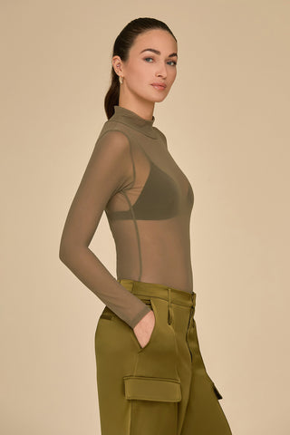 A person with long dark hair styled in a ponytail is wearing the elegant Giselle Bodysuit in olive mesh, paired with high-waisted olive green pants featuring pockets. They stand against a plain beige background with a confident expression.