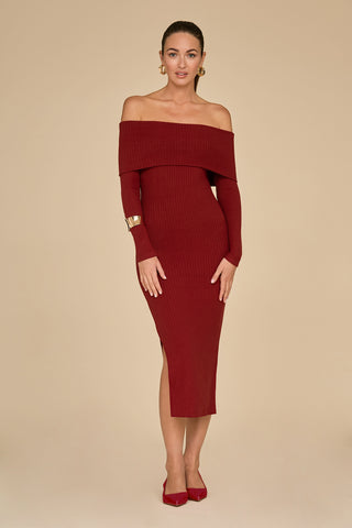 Against a beige background, a woman beams in a burgundy Amora Ribbed Knit Dress featuring an off-the-shoulder neckline and streamlined silhouette. Her ensemble is complete with gold hoop earrings, a bracelet, and red shoes. Her hair is pulled back elegantly, enhancing her poised presence.