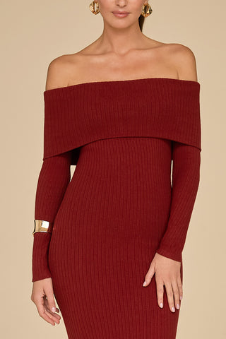 A person is wearing the Amora Ribbed Knit Dress in burgundy, showcasing an off-the-shoulder design with a streamlined silhouette and long sleeves. The ensemble is tastefully complemented by large gold hoop earrings and a gold cuff bracelet, all set against a solid neutral background.