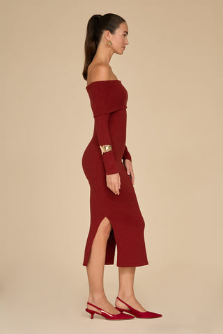 A woman stands in profile against a beige background, highlighting the Amora Ribbed Knit Dress - Burgundy with its off-the-shoulder design and streamlined silhouette. The long-sleeve, fitted burgundy dress features side slits, elegantly paired with red heels and gold accessories. Her hair is tied back in a ponytail.