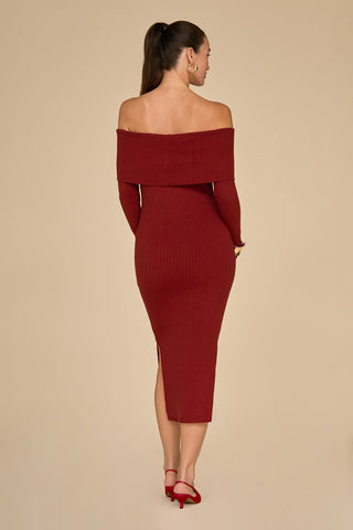 A woman in a streamlined silhouette, wearing an off-the-shoulder Amora Ribbed Knit Dress in burgundy, stands facing away. Her matching heels enhance her elegant look, while her hair is tied back in a ponytail. The plain beige background accentuates the striking ensemble.