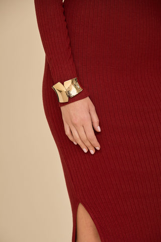 A person elegantly models the Amora Ribbed Knit Dress in burgundy, characterized by its high slit and streamlined silhouette. The dress boasts an off-the-shoulder design and is accessorized with a gold cuff bracelet on the wrist. The image highlights a hand resting on the hip against a neutral backdrop.