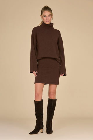 A woman is wearing the Calista Knit Skirt in Dark Oak, paired with a matching brown turtleneck sweater and knee-high black boots. She is standing against a beige background, smiling with her hair tied back.