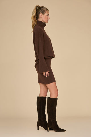 A woman wearing a dark oak turtleneck sweater and the matching Calista Knit Skirt pairs her outfit with dark brown knee-high boots. She stands sideways against a plain beige background, her hair tied back in a ponytail.