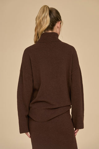 A person with long blonde hair in a ponytail is wearing the Calista Turtleneck Knit Sweater in Dark Oak. The person is standing with their back to the camera against a plain beige background.