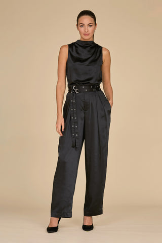 Against a beige background, an individual models a sleeveless black satin top paired with the Kora Double Belted Satin Pant in black by Noli Yoga. This stylish outfit is completed with black shoes, and their hair is neatly tied back.