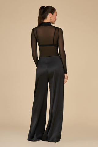 A woman with a ponytail stands with her back to the camera, wearing a black sheer long-sleeve top and Fleur Satin Pant - Black. The high-waisted, wide-leg design beautifully complements the subtle rose print against a plain beige background.