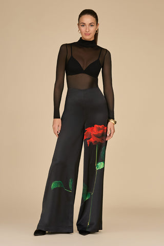 A woman stands confidently in a sheer black top and the Fleur Satin Pant in Black, adorned with a striking rose print. Her high-waisted, flare-leg pants complement her tied-back long hair and bracelet, all set against a neutral background.