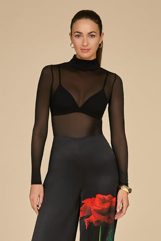 A woman stands against a beige background wearing the Giselle Bodysuit, a sheer black mesh turtleneck, paired with high-waisted black pants adorned with a red rose print. The high-neck design enhances her look, accompanied by her hair tied back, gold hoop earrings, and a watch on her left wrist.