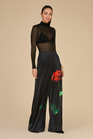 A woman stands against a neutral background wearing a black sheer top paired with the Fleur Satin Pant - Black, featuring an eye-catching rose print. The long flair of the pants displays a bold red rose design. Her hair is pulled back as she gives a slight smile.