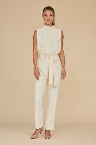 A person stands against a plain beige background, wearing the Charli Jumpsuit in Pearl—a sleeveless, high-neck cream ensemble with a tied waist. This statement piece is paired with nude heels and a gold bracelet, complemented by hair styled up and simple hoop earrings.