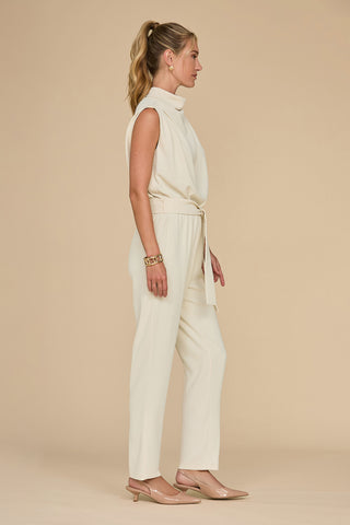 A woman wearing a Pearl Charli Jumpsuit is seen in profile against a beige background. This standout garment showcases a high collar and a waist tie, beautifully highlighting her gold bracelet and nude heels. Her hair is sleekly styled into a ponytail, enhancing the outfit's sophisticated appeal.