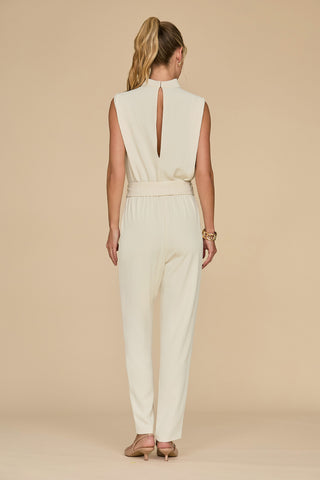 A person with a ponytail is wearing the Charli Jumpsuit - Pearl, a cream-colored sleeveless statement piece featuring a back slit and an elastic waistband. They are positioned on a beige background, facing away with one hand by their side and the other slightly bent.