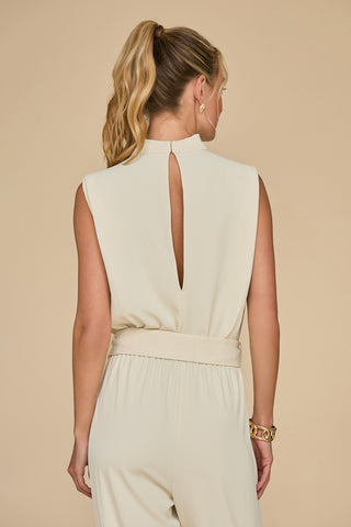 A woman with long blonde hair tied in a ponytail displays the Charli Jumpsuit - Pearl, a slim-fit statement piece featuring a keyhole back design. Set against a plain beige background, she accessorizes her chic ensemble with gold hoop earrings and a bracelet.