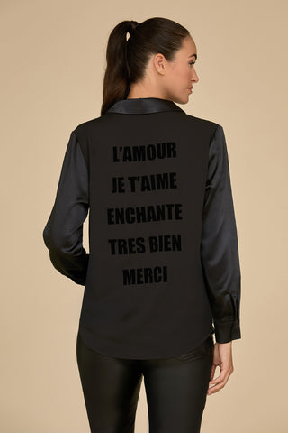 A woman with a ponytail is stylishly dressed in a chic statement piece—a L'Amour Silky Blouse in black adorned with French words such as "L'AMOUR," "JE T'AIME," and "ENCHANTÉ" printed on the back. She stands against a plain beige background, radiating effortless sophistication.