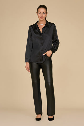 A person wearing the L'Amour Silky Blouse in black, paired with black leather pants and black heels, stands against a beige background. The blouse features a sheer mesh detail at the neckline, adding intrigue as they rest a hand in their pocket and showcase a necklace.