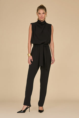 A woman stands against a beige background in a Charli Jumpsuit - Black, a sleeveless, slim-fit statement piece featuring a high collar and waist tie. She accessorizes with black heels and a gold bracelet, her hair elegantly styled up.