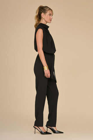 A woman in a slim-fit Charli Jumpsuit - Black stands in profile against a plain beige background. She has a blonde ponytail and wears black high heels and a gold bracelet, making her ensemble a true statement piece.