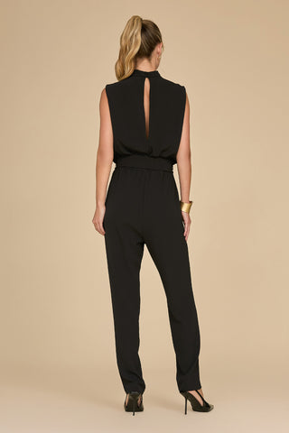 A person with long hair in a ponytail models the Charli Jumpsuit - Black, a chic, sleeveless statement piece, paired with high heels and accessorized with a gold bangle. The slim-fit jumpsuit showcases a keyhole back detail against a plain beige backdrop.