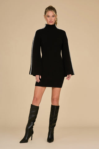 A person wearing the Raven Knit Dress in black and white, featuring long sleeves and white detailing along the edges, paired with knee-high black boots. The background is a plain beige color.