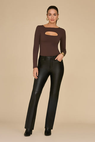 A woman poses against a beige background, wearing the Jada Cut Out Long Sleeve Top in espresso and black leather pants. Her hair is pulled back, and she accessorizes with a bracelet and earrings. The curve-flattering material of the top enhances her confident stance and neutral expression.