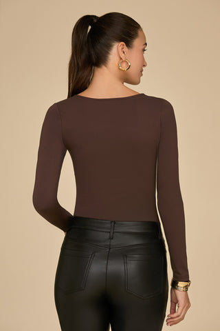 A woman with a ponytail wears the Jada Cut Out Long Sleeve Top in espresso, crafted from curve-flattering material, paired with black leather pants. She accessorizes with gold hoop earrings and a gold wristwatch, her back turned to the camera against a beige background.