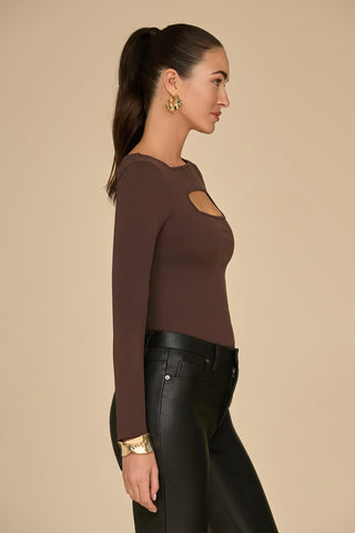 A woman with long dark hair tied back wears the Jada Cut Out Long Sleeve Top in Espresso, a fitted piece with curve-flattering material and keyhole cutout, paired with black leather pants. She accessorizes with large gold earrings and a gold bracelet, standing against a plain beige background.