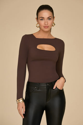 A person with long hair styled up, showcasing gold hoop earrings and a bracelet, is dressed in the flattering Jada Cut Out Long Sleeve Top in espresso. They pair it with black leather pants, standing elegantly against a plain beige background.