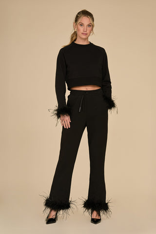 A woman stands confidently against a beige background, wearing a Rada Feather Trim Crewneck in black, paired with black pants featuring ostrich feather trim at the wrists and ankles. Complemented by chic black high heels, her hair is elegantly tied back in a ponytail.