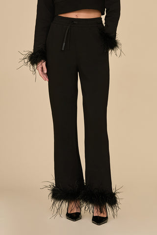 A person is dressed in a chic outfit, showcasing the Rada Feather Trim Pant - Black with ostrich feather accents on the sleeves and pant legs. The wide-legged design flows gracefully as they pose against a beige background, highlighted by sleek black pointed shoes.