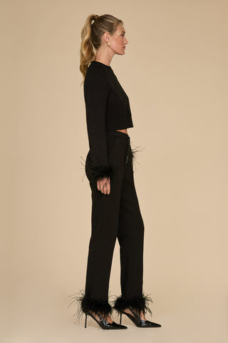 A woman with blonde hair in a ponytail stands sideways, elegantly dressed in the Rada Feather Trim Pant outfit in black, crafted from French Terry material and embellished with ostrich feather trim on the sleeves and pant legs. She enhances her chic look with black pointed heels against a beige background.