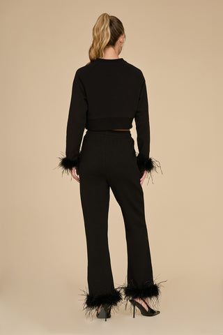 A person faces away from the camera, dressed in a black two-piece ensemble with ostrich feather trim on the sleeves and wearing the Rada Feather Trim Pant - Black. Their long hair is styled in a high ponytail, and they are sporting black high heels against a plain beige background.