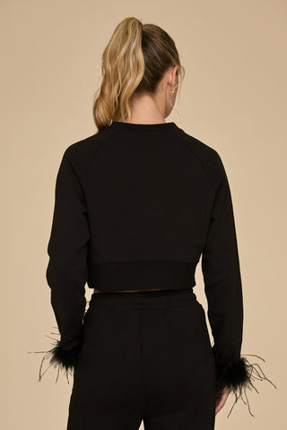 Standing against a beige background, an individual with long blonde hair styled in a ponytail gracefully wears the Rada Feather Trim Crewneck in Black, paired with coordinating pants adorned with ostrich feather trim on the sleeves as they face away from the camera.