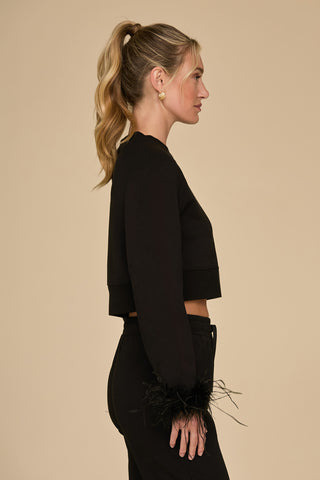 A woman in a stylish black Rada Feather Trim Crewneck poses in profile against a beige background. Her long blonde hair is pulled back into a ponytail, and she accessorizes with gold earrings.