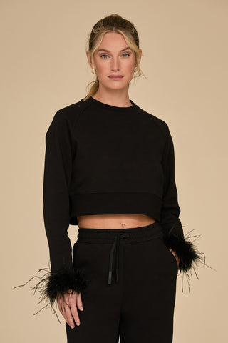 A person with long hair is wearing a black Rada Feather Trim Crewneck that features ostrich feather trim on the cuffs, paired with matching black pants. The background is a plain beige color, highlighting the elegant ensemble.