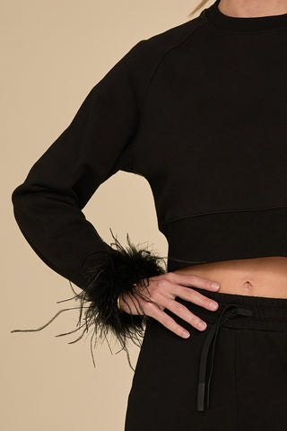 Wearing the elegant Rada Feather Trim Crewneck in black, featuring an ostrich feather detail on the cuffs, a person pairs it with black jogger pants. Striking a pose with one hand on their hip, they confidently present the ensemble against a plain beige background.