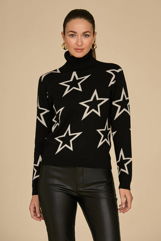 A person is wearing the Astra Ski Sweater in black, featuring white star patterns, paired with black pants. They stand against a beige background, with their long hair pulled back and large hoop earrings completing the look.