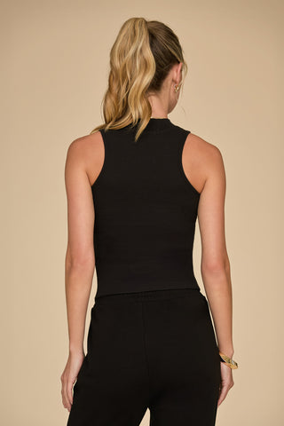 A person with long blonde hair tied in a ponytail is facing away from the camera, wearing the Talia Ribbed Tank - Black and black pants. The ultra-soft rib fabric of the top adds texture against the neutral beige background.