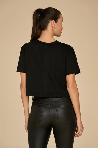 A person with long, brown hair styled in a ponytail is facing away, wearing a black cropped fit t-shirt that resembles the Naya Suede T-Shirt - Black and paired with black leather pants. The plain beige background accentuates the ensemble as a chic layering piece.